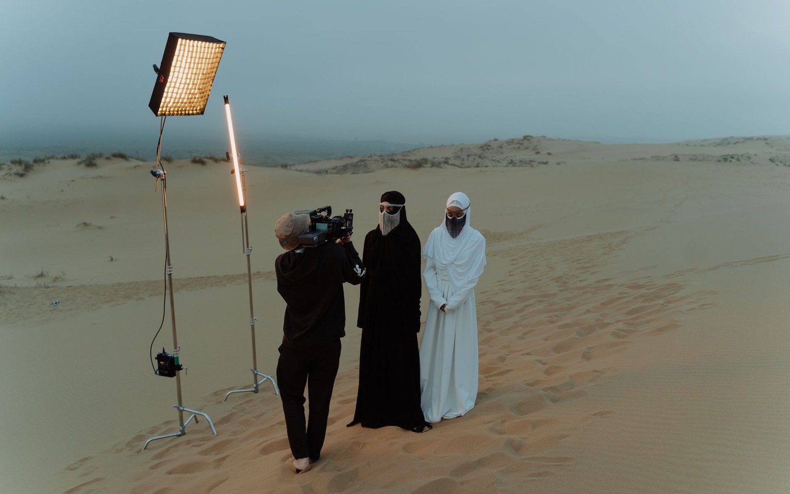 Film crew at work on a desert dolly track — camera operator at the eyepiece, focus puller and dolly grip beside him, dunes behind, all adults