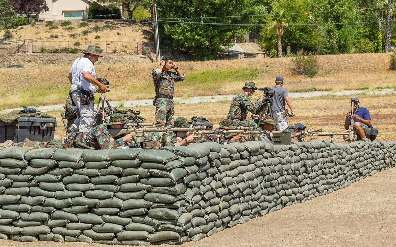 Snipers in camouflage take firing positions behind a sandbag barrier at an outdoor range