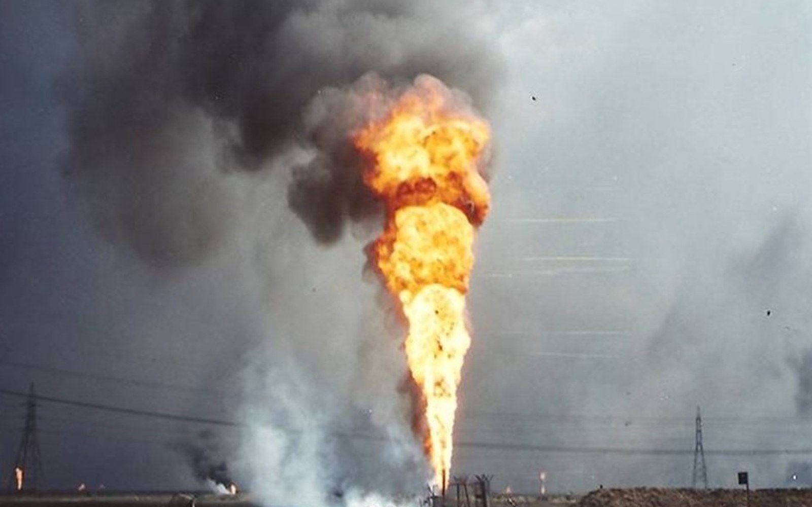 A tall fire and thick black smoke shoot skyward from a burning oil well in an open field