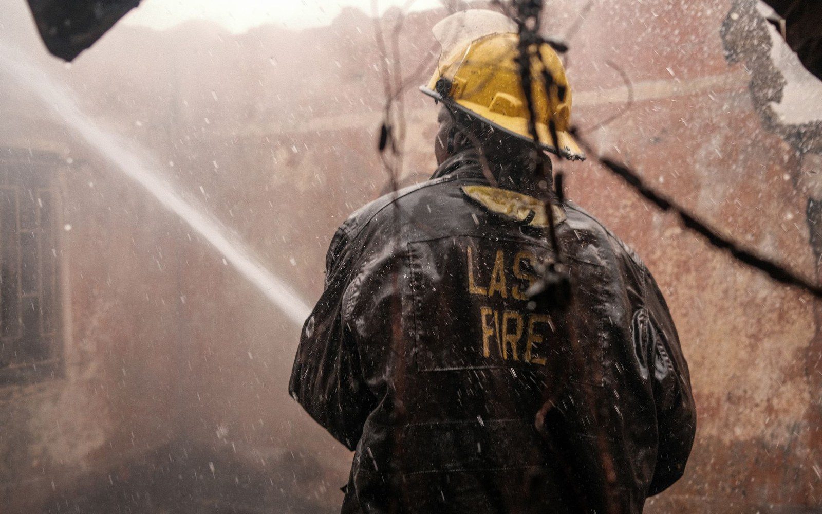 A firefighter in a yellow helmet sprays water at a smoldering, burned-out building