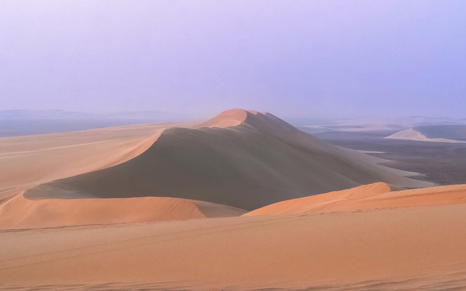 Sweeping sand dunes cast in shadow under a hazy desert sky