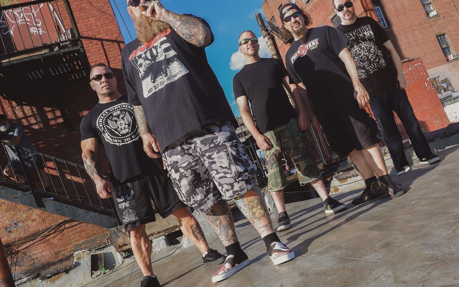 Five men in black t-shirts and shorts stand on a rooftop between brick buildings