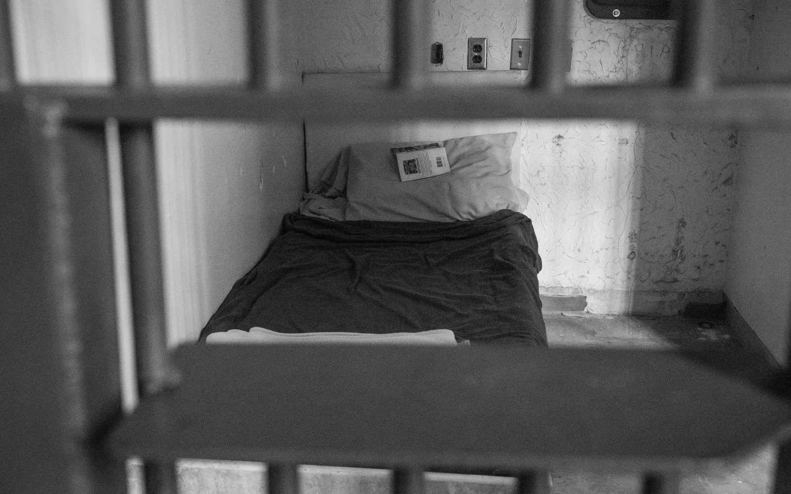 Bars frame a narrow jail cell bed with a folded blanket and pillow