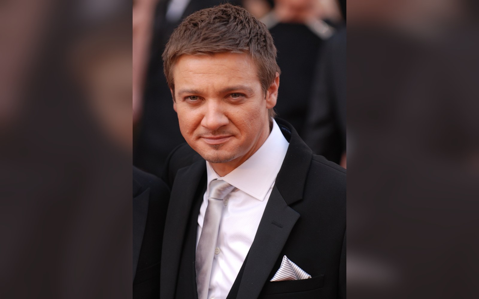 Man in a black tuxedo with white shirt and tie smiling on a red carpet