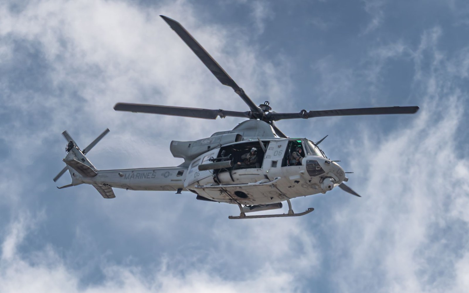 A US Marines helicopter flies against a cloudy sky with visible pilots aboard