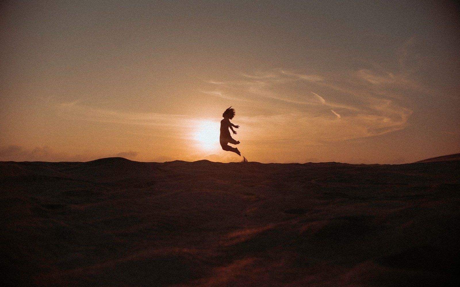 Silhouette of a person leaping mid-air against a glowing sunset over dunes