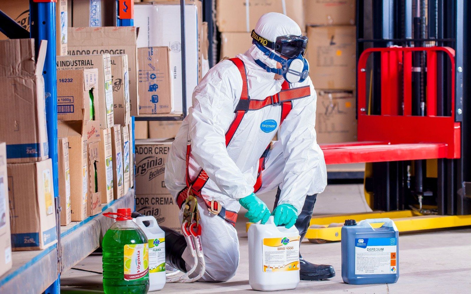 Worker in a protective suit and respirator kneels to inspect chemical containers in a warehouse