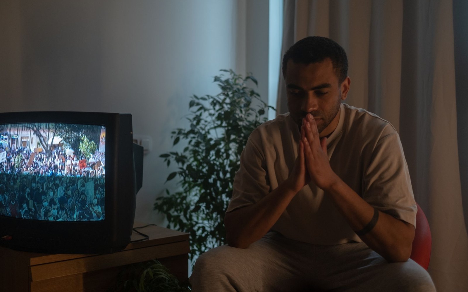 A man sits with hands clasped in prayer while a crowded protest plays on an old television